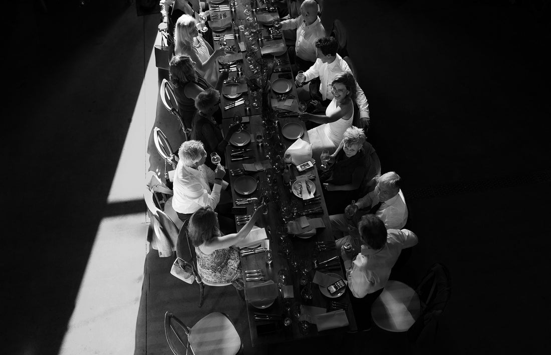 Black and white photo of people sitting around a long dining table, engaged in conversation and holding drinks; sunlight streams onto part of the table, creating dramatic shadows—a perfect moment for a Creative Agency for Wine  Hospitality to capture and elevate.