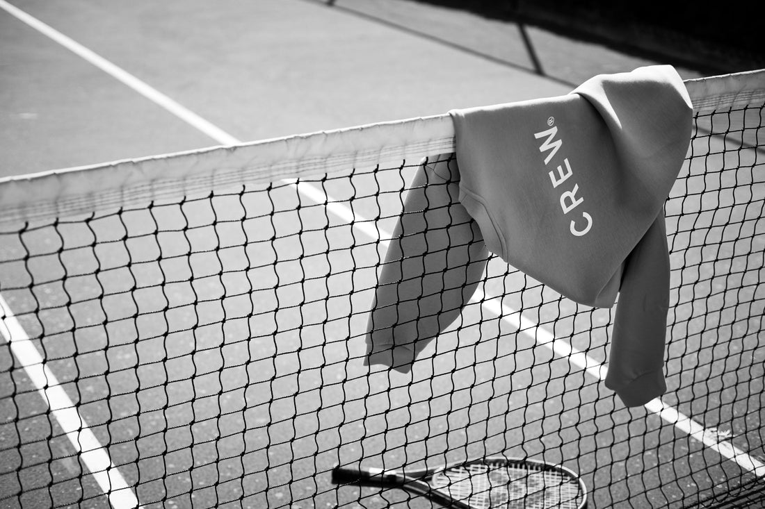 A tennis sweatshirt labeled CREW hangs over a net on an outdoor tennis court, with a tennis racket lying on the ground below. The black and white image captures the refined touch of a Hospitality Branding Agency.