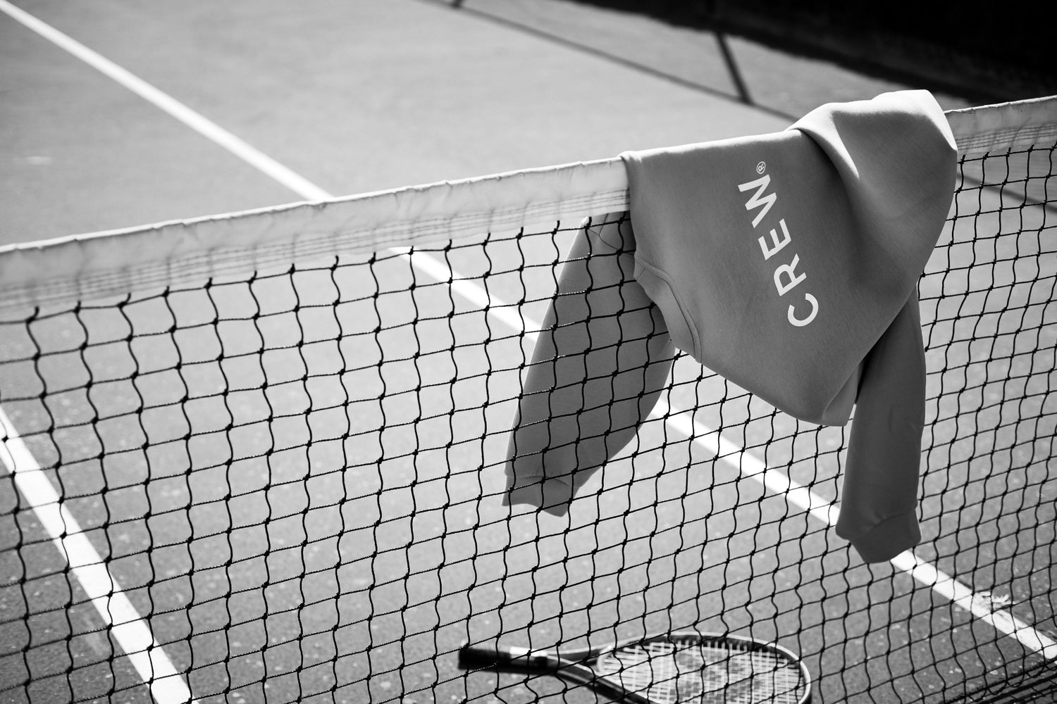 A tennis sweatshirt labeled CREW hangs over a net on an outdoor tennis court, with a tennis racket lying on the ground below. The black and white image captures the refined touch of a Hospitality Branding Agency.