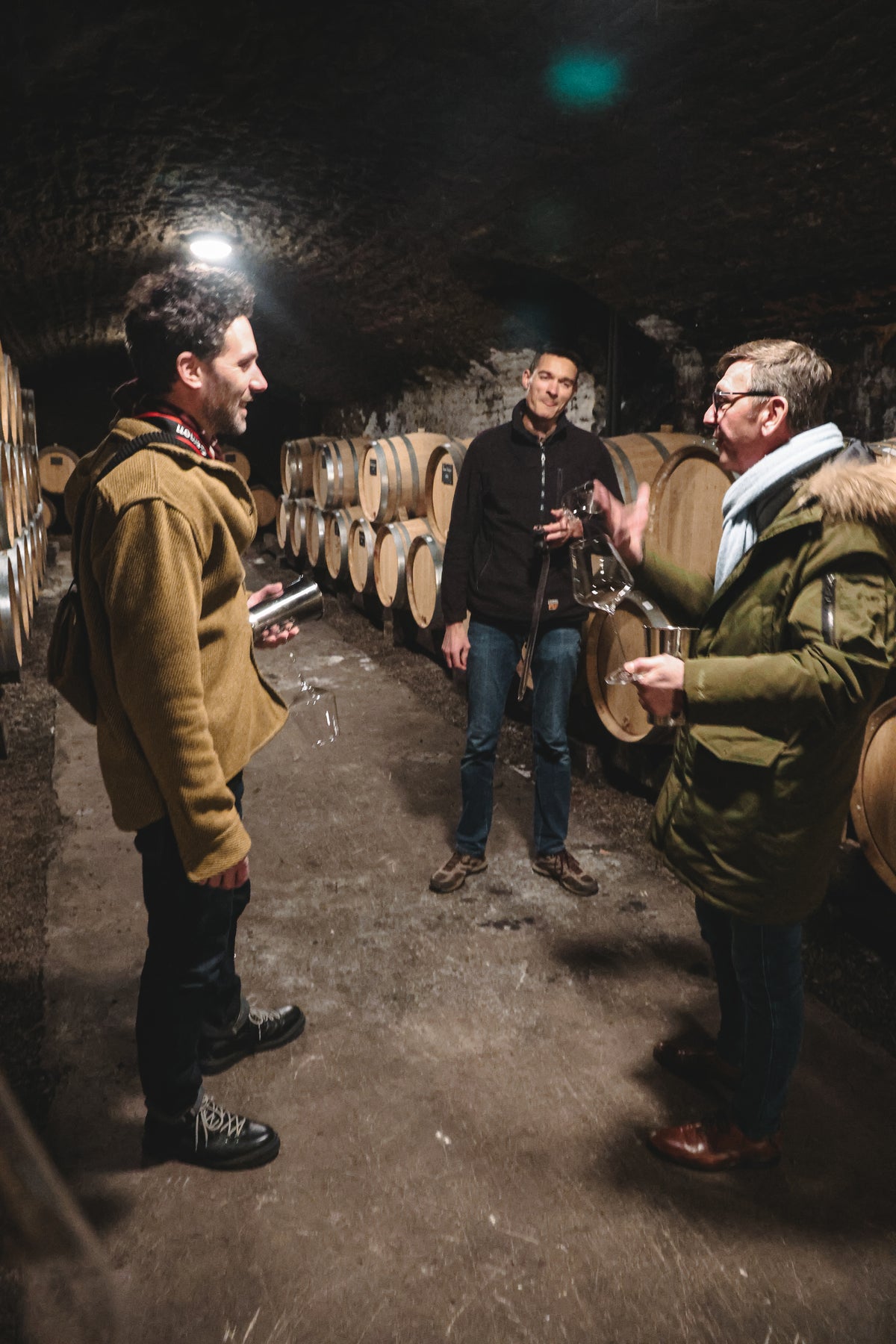 Three people stand talking in a dimly lit wine cellar lined with wooden barrels. One gestures with a glass while others listen, empty glasses in hand—a perfect setting for a hospitality branding agency specializing in wine and branded merchandise for wineries.