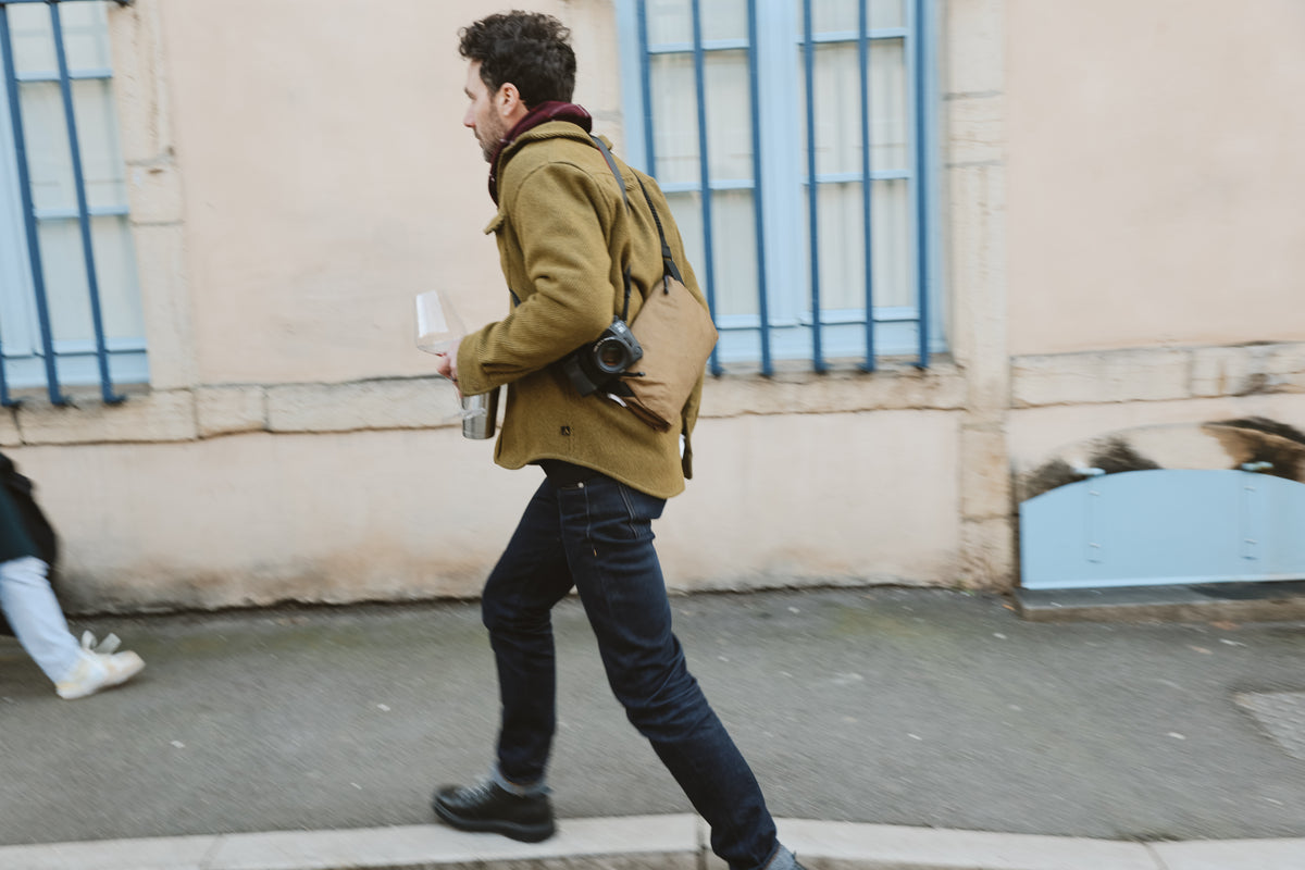A man wearing a green coat, jeans, and a brown bag walks briskly along a sidewalk, carrying a drink and camera—perhaps scouting inspiration for a Hospitality Branding Agency—with blue window frames on a beige building in the background.