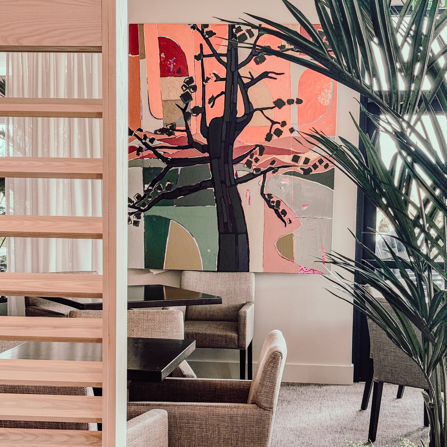 A modern lounge area with beige chairs and black tables, perfect for a Creative Agency for Wine  Hospitality, features a colorful abstract tree painting and large green plants. Light wood slats partially divide the inviting space.