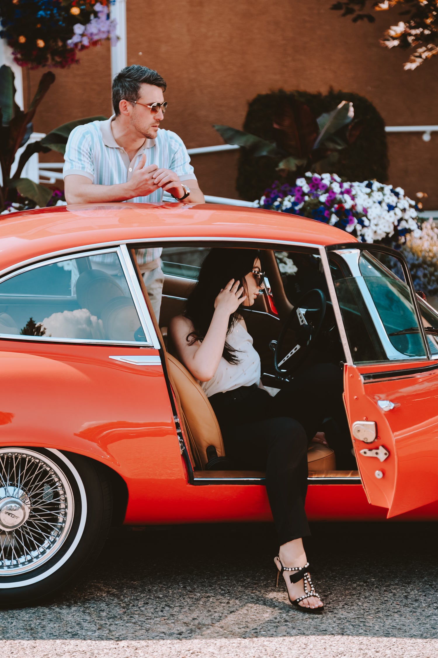 A man leans on the roof of a vintage red car while a woman in sunglasses and heels sits in the driver’s seat, adjusting her hair. Colorful flowers bloom in the background—perfect inspiration for creative agency wine label  packaging design.