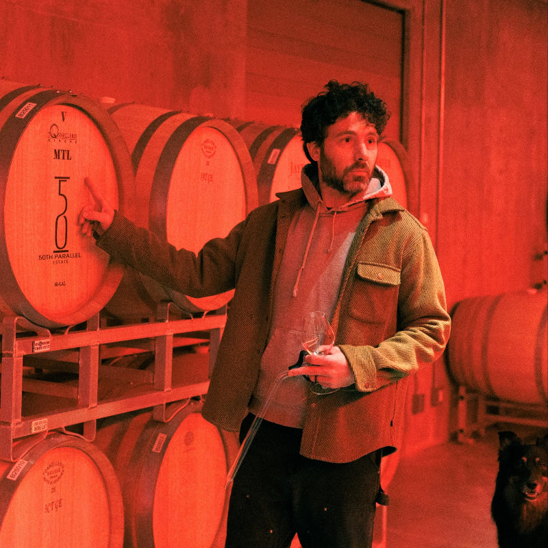 A man holding a wine glass and a wine thief stands in a dim red-lit cellar, pointing at a wooden wine barrel. Stacks of barrels line the walls—a scene perfect for inspiration from a Creative Agency for Wine  Hospitality. A black dog is partially visible nearby.