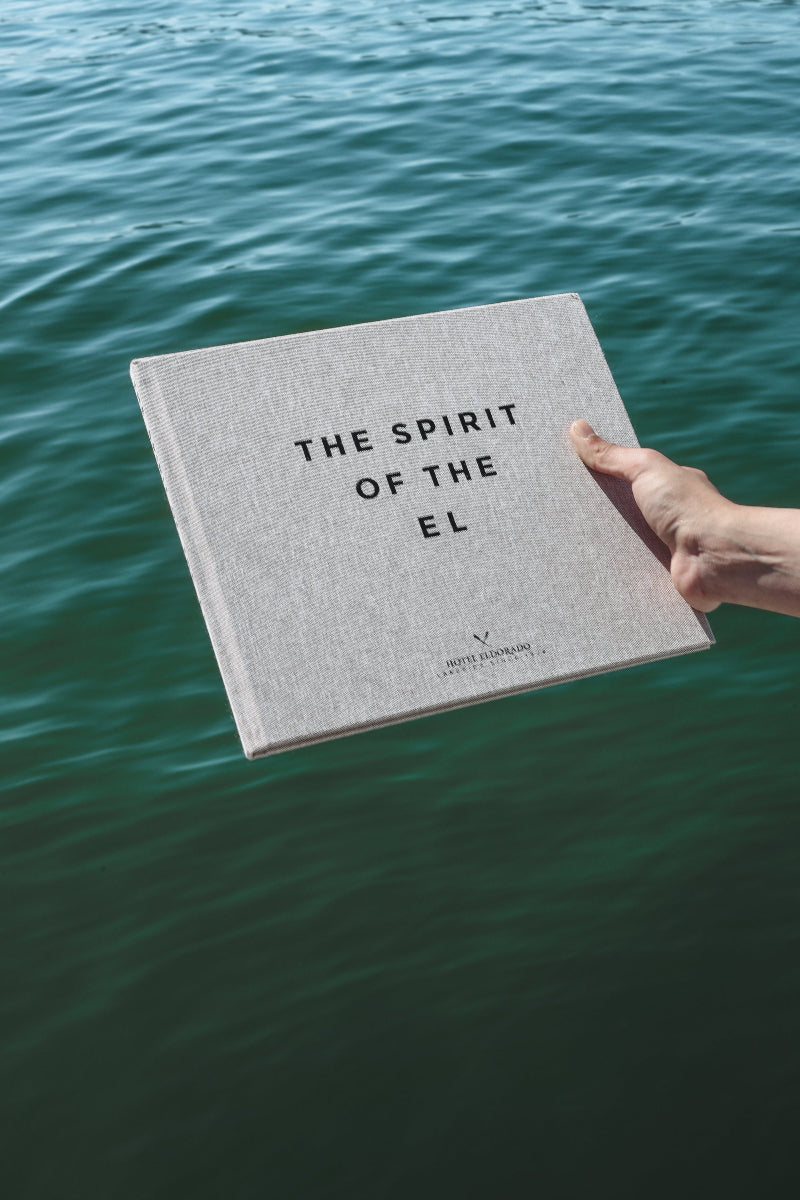 A hand holds a beige book titled THE SPIRIT OF THE EL, echoing the refined touch of hospitality branding boutique hotel identity, over a body of rippling blue-green water. 