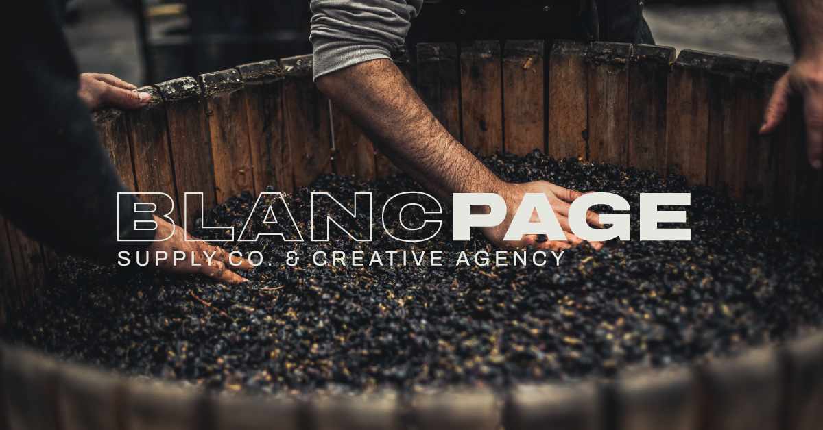Two people’s hands mix or crush dark grapes in a large wooden vat. Overlaid text reads “BLANC PAGE SUPPLY CO.  CREATIVE AGENCY.” The scene suggests traditional winemaking by a creative agency for wine  hospitality.