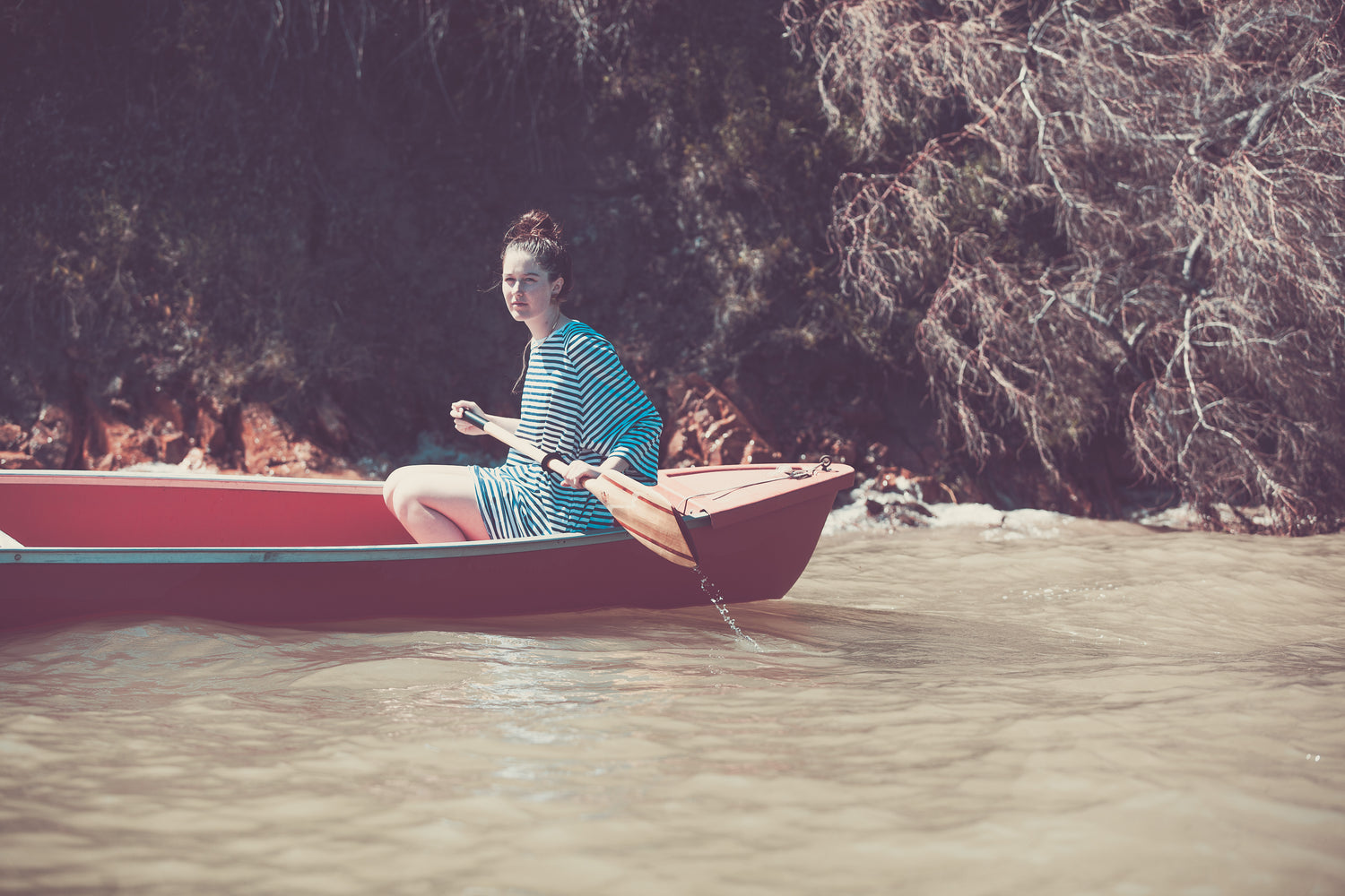A person in a striped dress sits in a red rowboat, holding an oar on muddy water near a wooded, rocky shoreline. Sunlight illuminates the scene, perfect inspiration for wine label  packaging design by a hospitality branding agency.