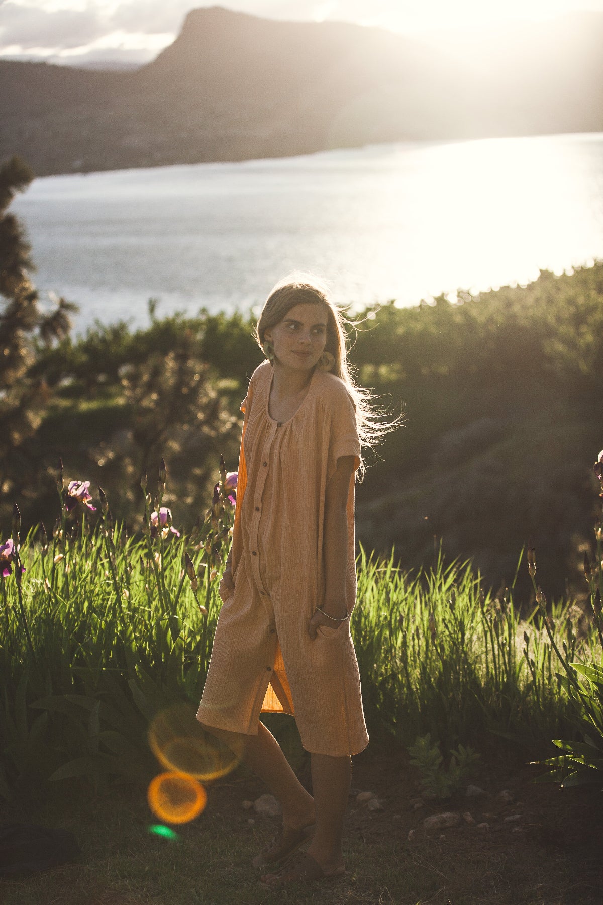 Woman in a peach dress standing in a field with a lake and mountains in the background. beautiful image for an airnbnb host showing off the Okanagan.