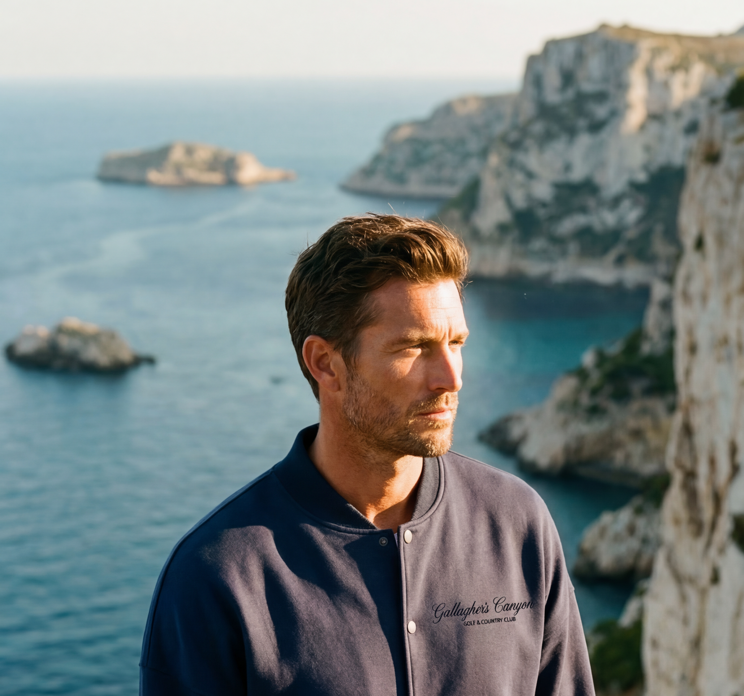 A man with short brown hair and a navy jacket stands on a cliffside, gazing into the distance—an inspiring scene that could grace the portfolio of a Creative Agency for Wine  Hospitality. Rocky cliffs and blue ocean waters complete the backdrop.