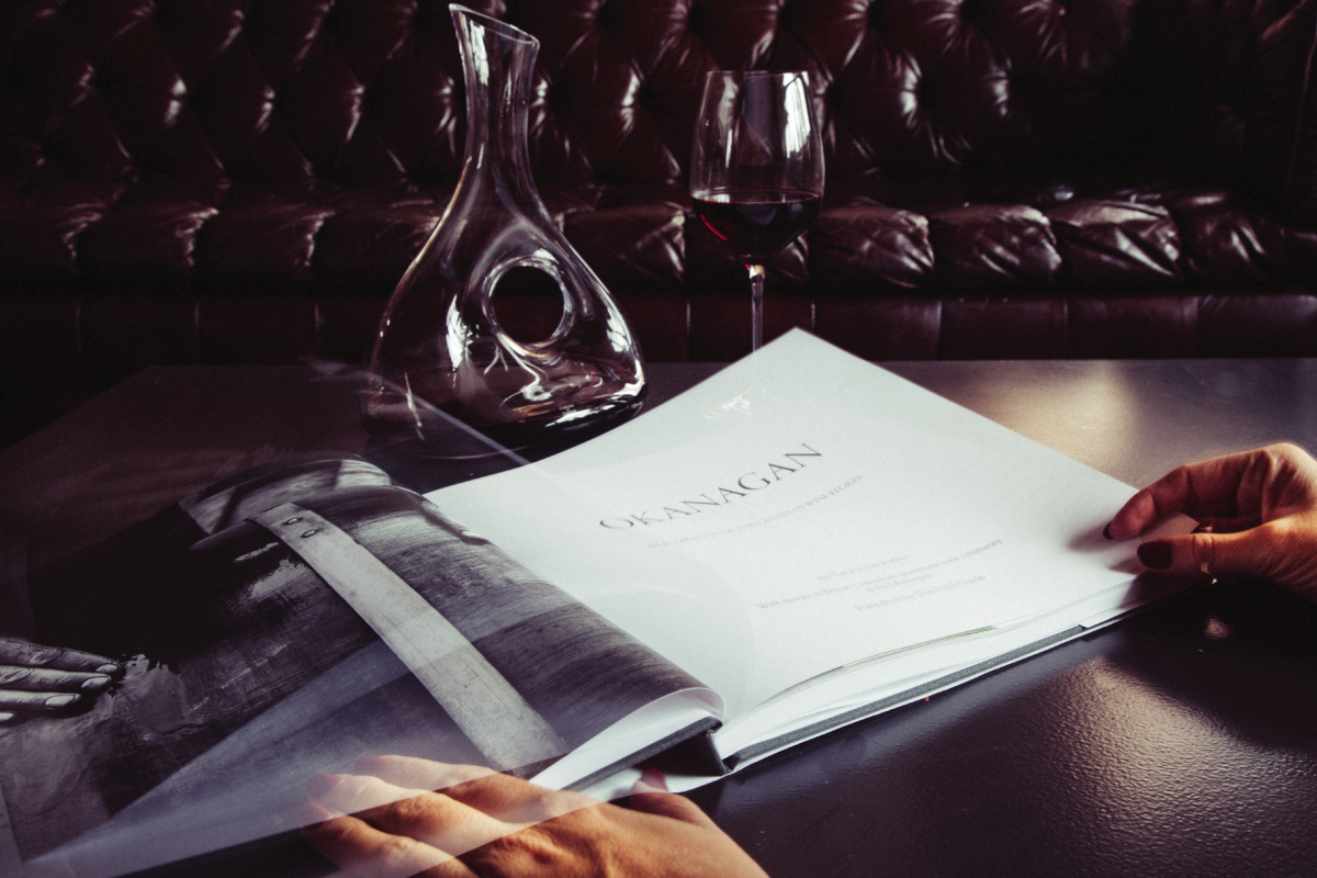A person’s hands hold open a large book titled “OKANAGAN” on a table with a wine decanter and a glass of red wine—an inspiring setting for anyone interested in wine label  packaging design or branded merchandise for wineries.