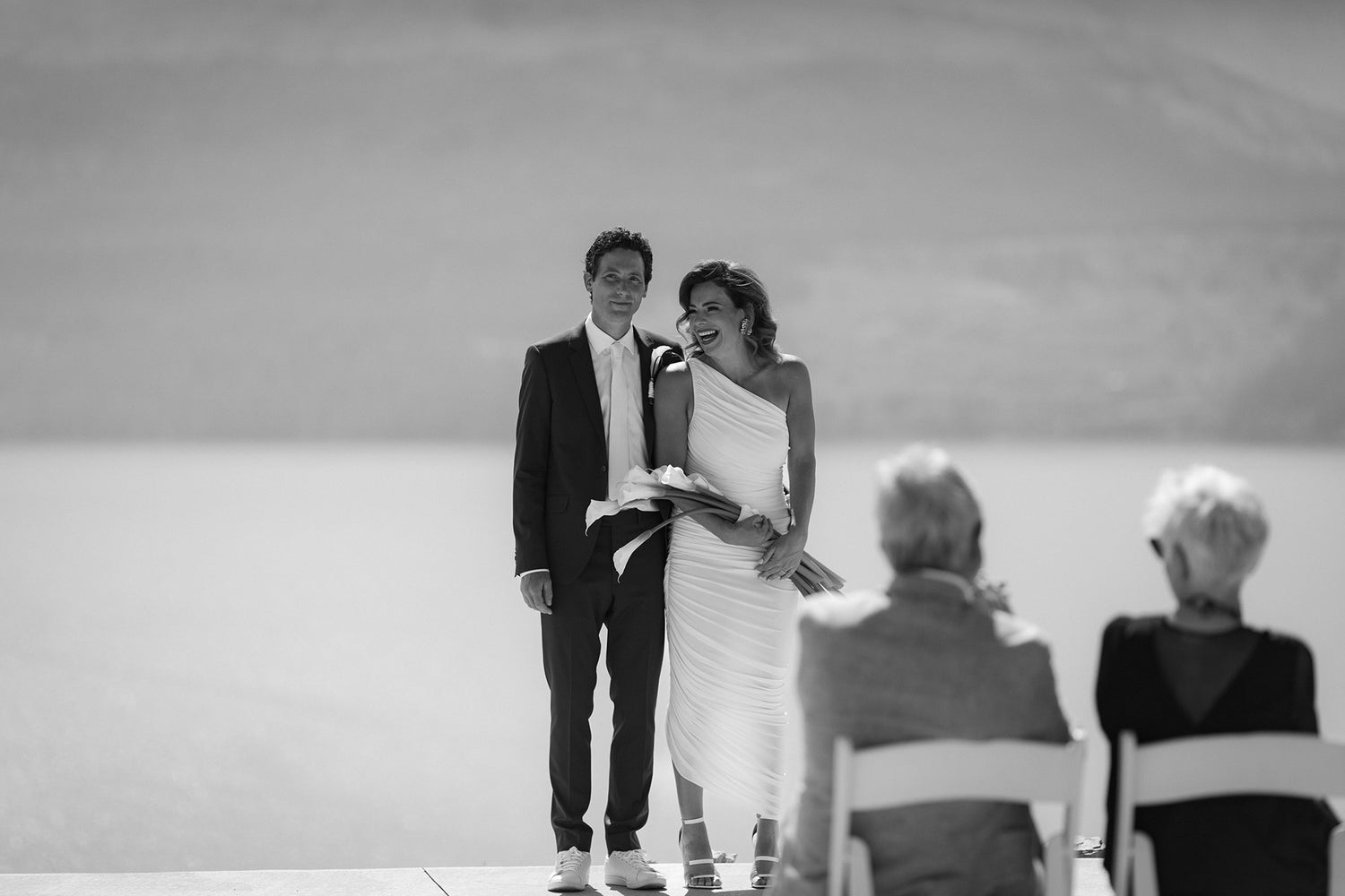 A couple stands together at an outdoor wedding ceremony; the woman in a white dress holds flowers and leans happily on the man in a suit, while two guests watch. The serene backdrop hints at expert Wine Label  Packaging Design by a top Winery Branding Agency.