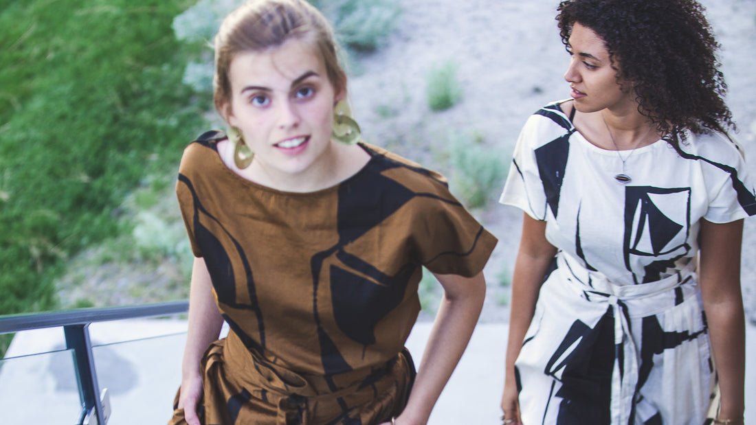 Two women in patterned dresses walk up outdoor stairs; one smiles at the camera while the other looks toward her. Greenery and a glass railing set the scene—perfect for a winery branding agency’s creative showcase.