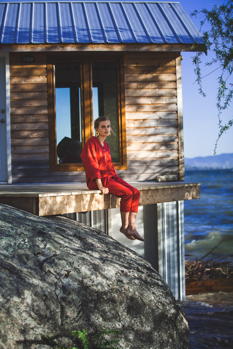 A person in a red outfit sits on a wooden cabin’s deck overlooking water and rocks, sunlight casting shadows as trees frame the scene—an inspiring view for any hospitality branding agency or airbnb host seeking natural elegance.