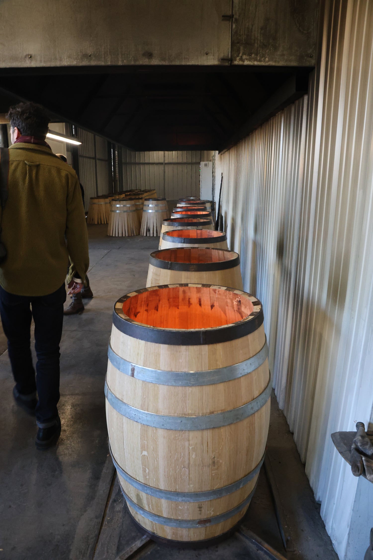 A row of wooden barrels with glowing orange interiors, possibly from charring, lined up inside a metal-walled facility. A person in a green jacket walks past—evoking the artistry of a Creative Agency for Wine  Hospitality.