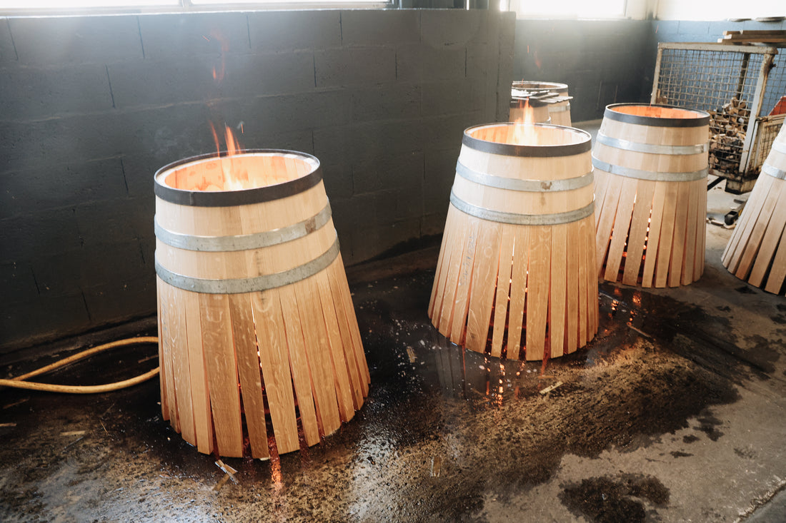 Three wooden barrels being charred with fire inside a workshop, perfect for a Winery Branding Agency. The barrels are unfinished, with metal hoops and open bottoms, flames flickering inside each one. The wet floor and dark wall create an atmospheric scene.