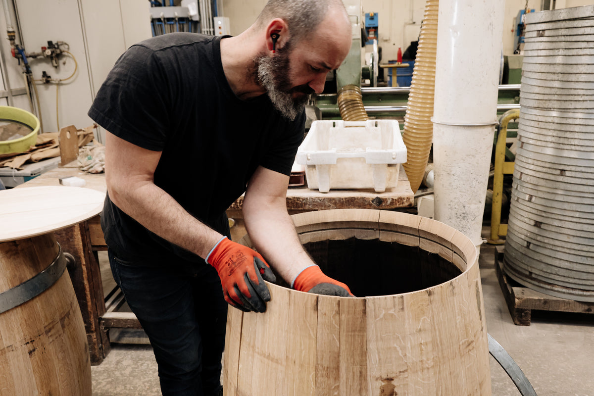 A man wearing red gloves and a black shirt assembles a wooden barrel in a workshop filled with tools—an authentic scene reflecting the craftsmanship behind branded merchandise for wineries.