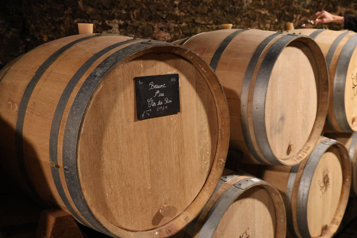 Several wooden wine barrels are stacked in a dimly lit cellar, evoking timeless charm. A small label reads “Beaune, Clos du Roi, 2021.” The rustic setting is ideal inspiration for any creative agency for wine  hospitality.