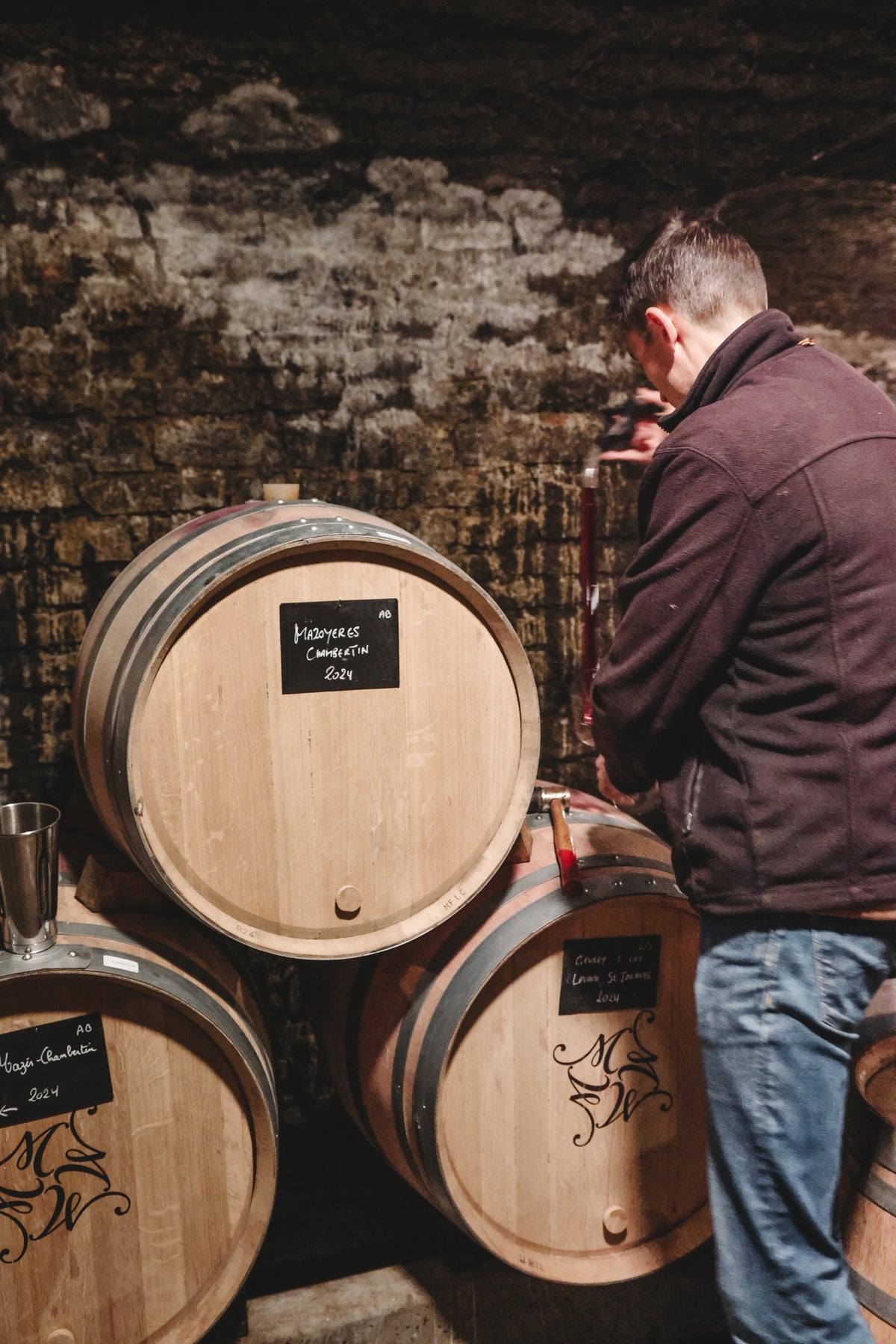 A man in a dark jacket samples wine from a wooden barrel in a dimly lit cellar, stacked with oak barrels—a perfect scene for a Winery Branding Agency capturing authentic moments for wine label  packaging design.