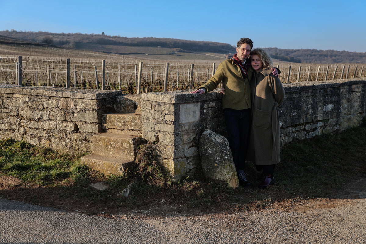 A man and woman in coats stand close together, smiling by a stone wall and steps in a rural vineyard—an inviting scene that captures the warmth celebrated by any Creative Agency for Wine  Hospitality under a clear blue sky.