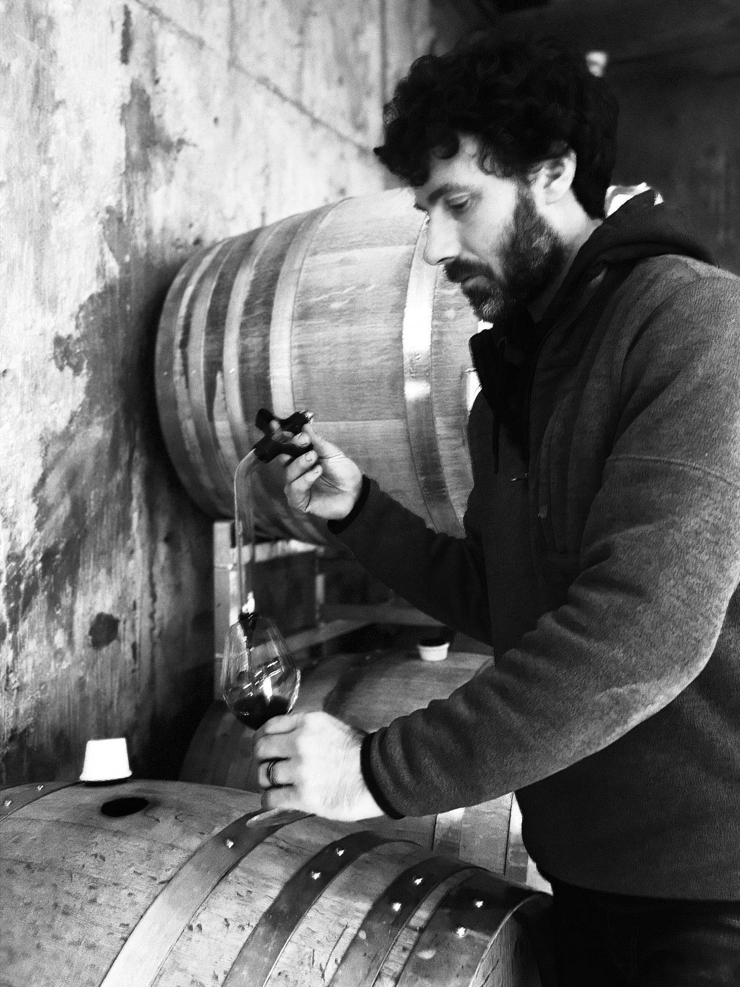 A man stands next to wooden barrels in a winery, using a wine thief to pour red wine from a barrel into a glass. The scene, captured in black and white, highlights the artistry behind wineries and their Wine Label  Packaging Design.