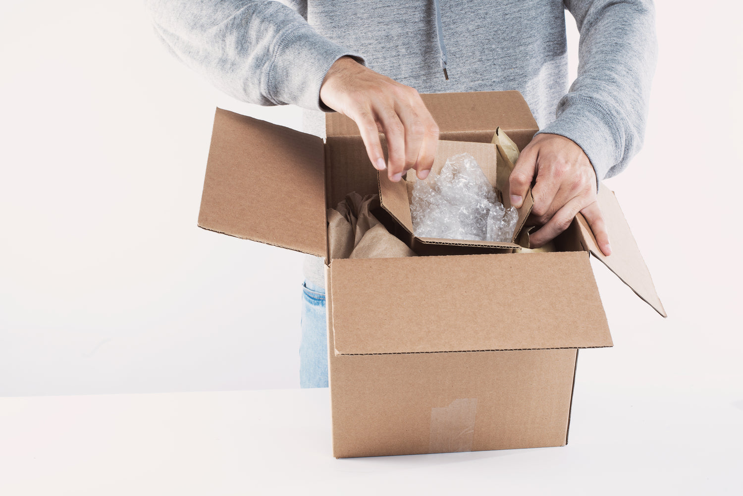 A person in a gray hoodie is packing bubble-wrapped items into a cardboard box, ideal for Wine Label  Packaging Design or branded merchandise for wineries, on a white surface with a clean white background.